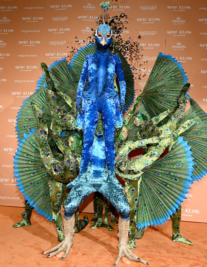 Heidi Klum in a detailed blue peacock costume with large feathered fans and dancers in matching green peacock outfits.