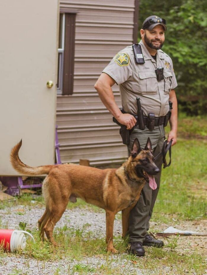 Police officer standing outdoors with a K9 unit dog near a building in a rural area.