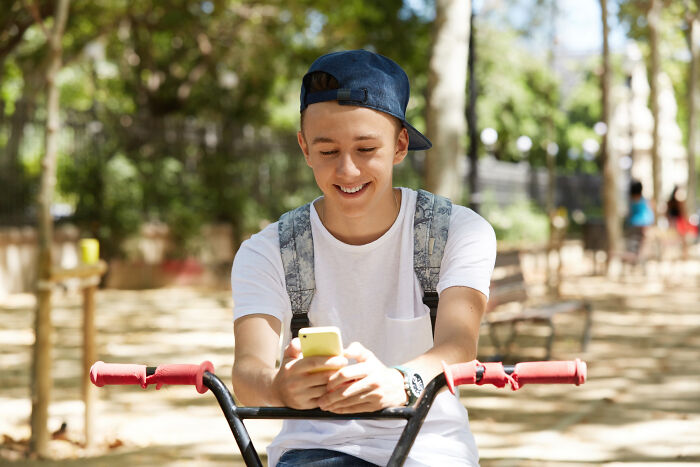 Teen boy wearing a cap and backpack on a bike, smiling while looking at his phone, showcasing misreading social cues.