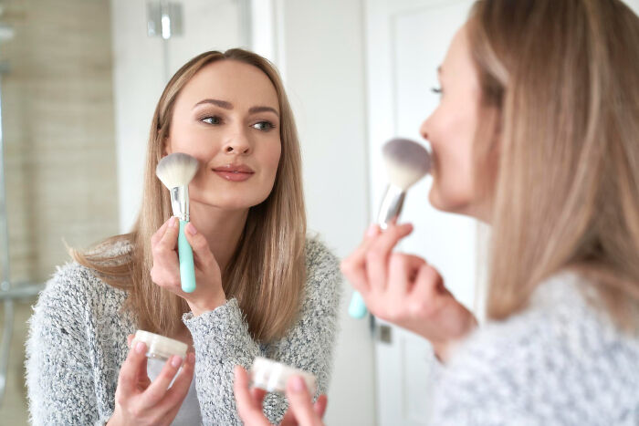 Young woman applying makeup with brush in front of mirror, reflecting confidence and focus on her routine.