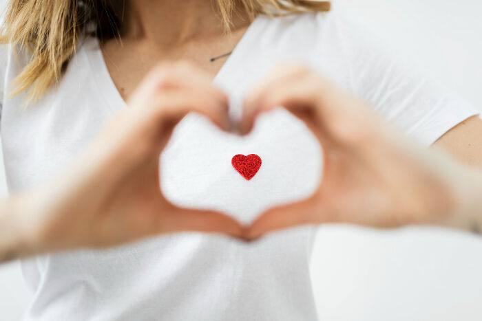 Person forming a heart shape with hands over chest wearing white shirt with a small red heart symbol in the center.