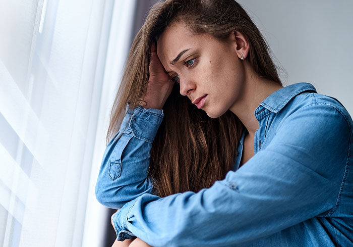Sad woman in blue shirt looking out window, reflecting on a man who proposed after being dumped two months earlier.