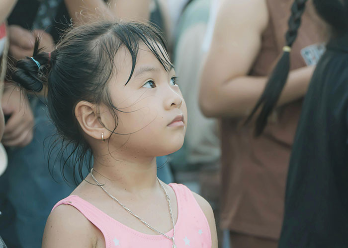 Young girl with a pink top looking up thoughtfully among a crowd, capturing kids embarrassing moms moments.