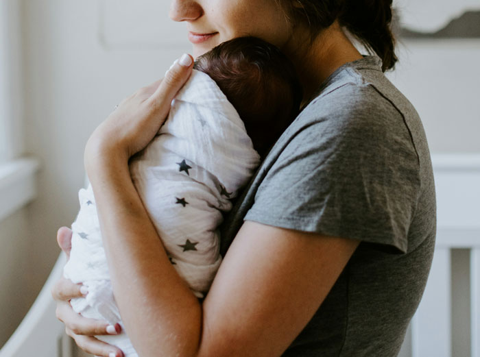 Woman in gray shirt gently holding a swaddled baby, illustrating humorous moments of unaware common knowledge.