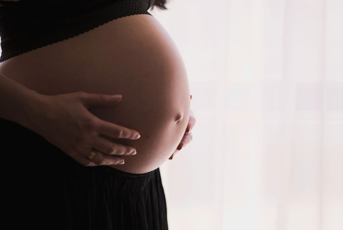 Pregnant woman gently holding her belly near a window, illustrating themes related to daycare workers and family secrets.