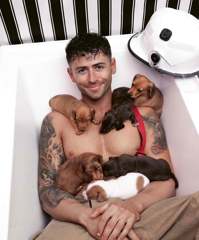 Firefighter with tattoos smiling in a bathtub surrounded by adorable puppies, firefighter helmet nearby for Australian calendar.