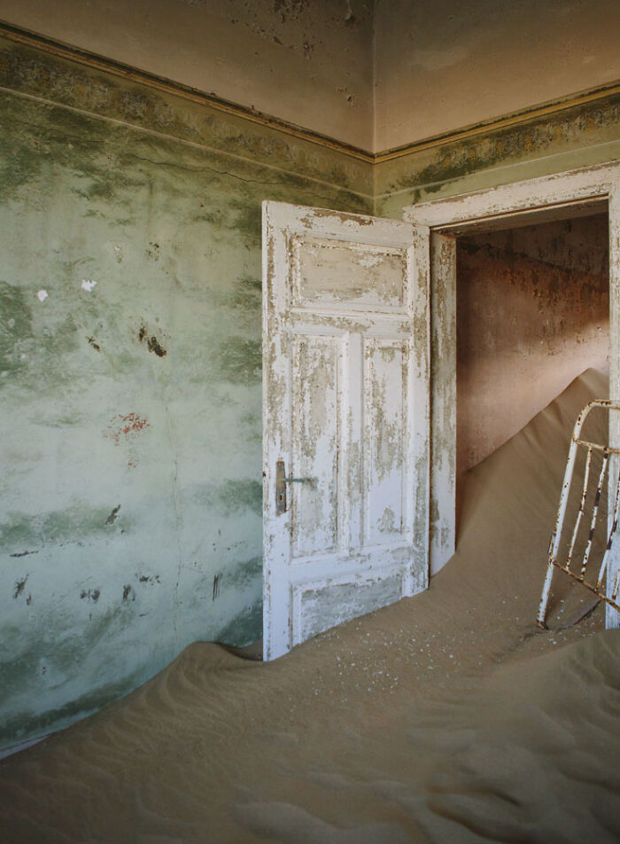 Abandoned home interior with peeling paint and desert sand slowly swallowing the floor through an open door.
