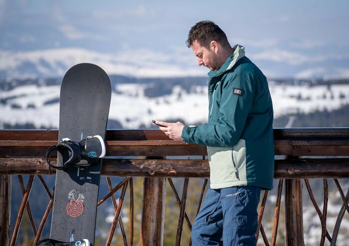 Man standing next to snowboard on wooden railing, looking at phone, showcasing lovable clueless dads during pregnancy moments.