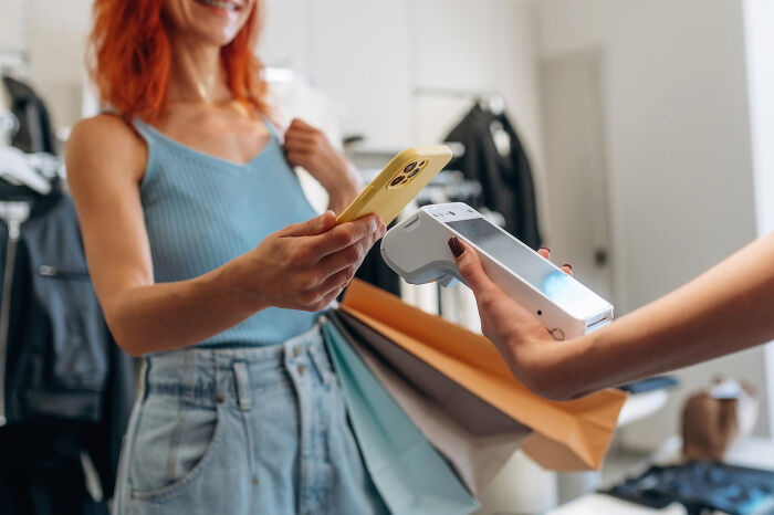 Woman using smartphone to make a contactless payment while holding shopping bags, illustrating clever life hacks and extra work realization.