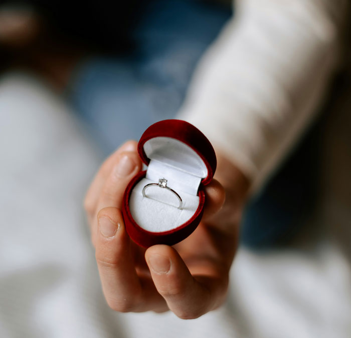 Hand holding red ring box with engagement ring, symbolizing man proposes two months after girlfriend dumps him.