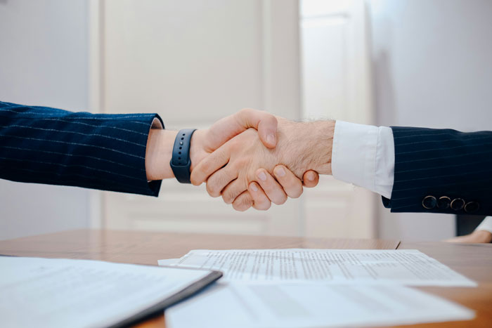 Two professionals shaking hands over a desk with documents, illustrating company claims about raises and new hire salary.