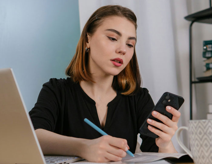 Young woman in black top working remotely, looking at phone and writing notes about unexpected work situations.