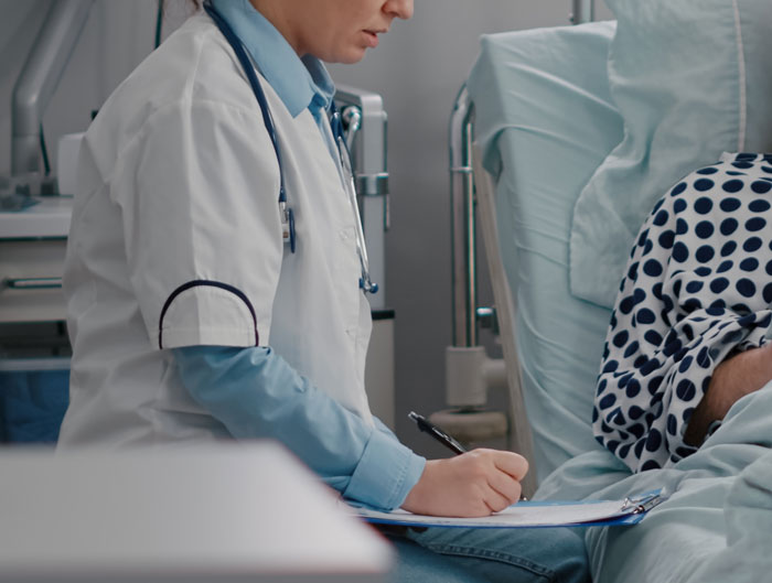 Doctor in white coat taking notes while sitting next to a patient in a hospital bed, showing cruel person contrast.