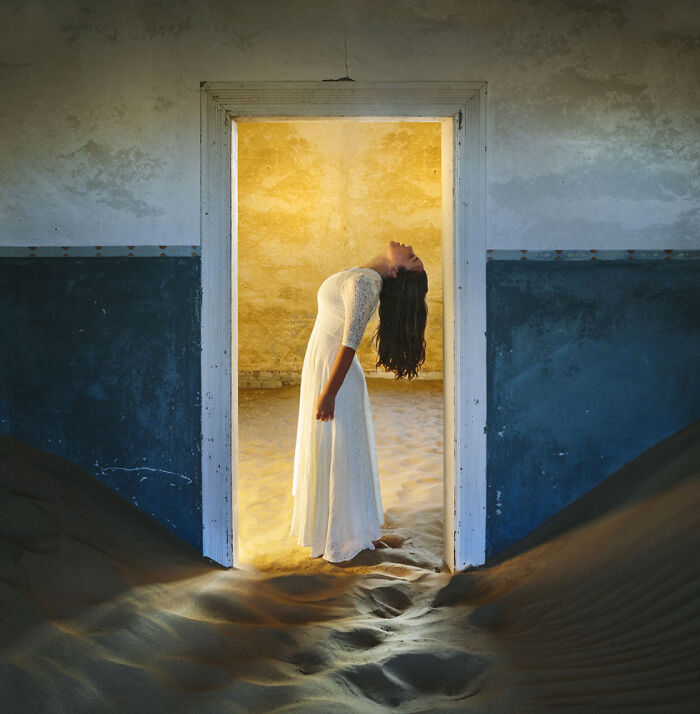 Woman in a white dress leaning back inside an abandoned home partially filled with desert sand dunes.