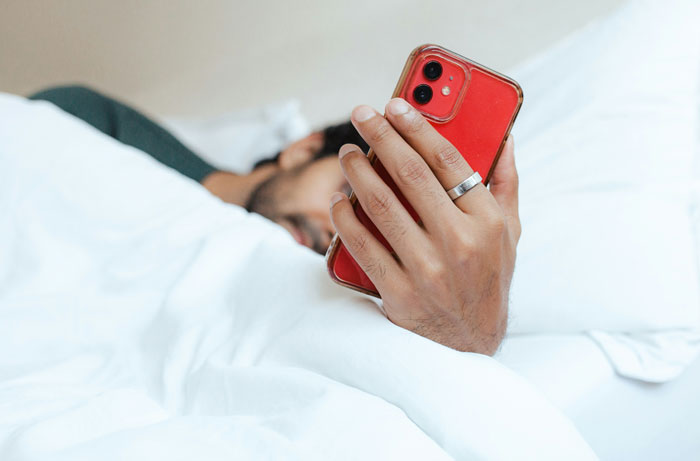Man lying in bed under white sheets holding a red smartphone, illustrating one-in-a-million coincidences concept.