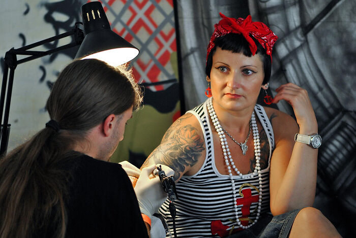 Tattoo artist working on a detailed sleeve tattoo for a woman with a red bandana and nautical-themed accessories.