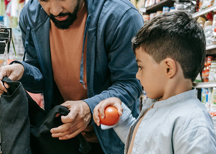 A father and son grocery shopping together, with the child holding a tomato in a store aisle filled with products.