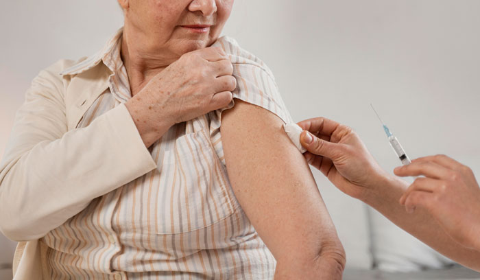 Elderly woman receiving a vaccine injection in her upper arm from a healthcare professional indoors.