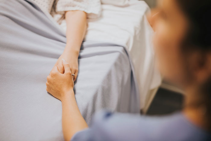 Person holding a patient’s hand in a hospital bed, highlighting trust in plastic surgery consultations.