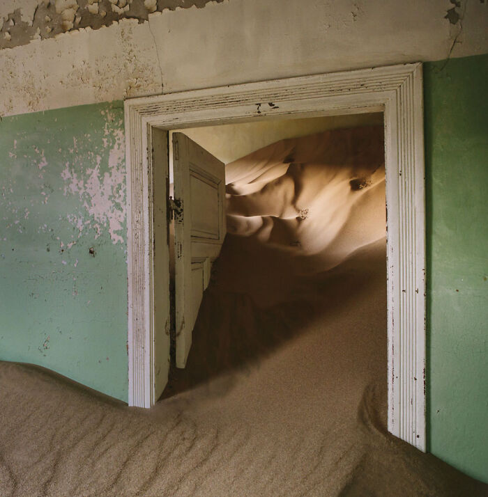 Desert sand filling abandoned home interior, creating surreal sand castles and dunes inside old doorframe.