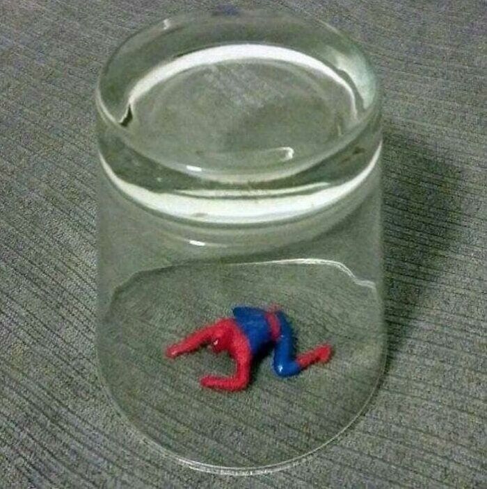 Tiny Spider-Man figure appears trapped under a glass on a textured gray surface in a confusing image.