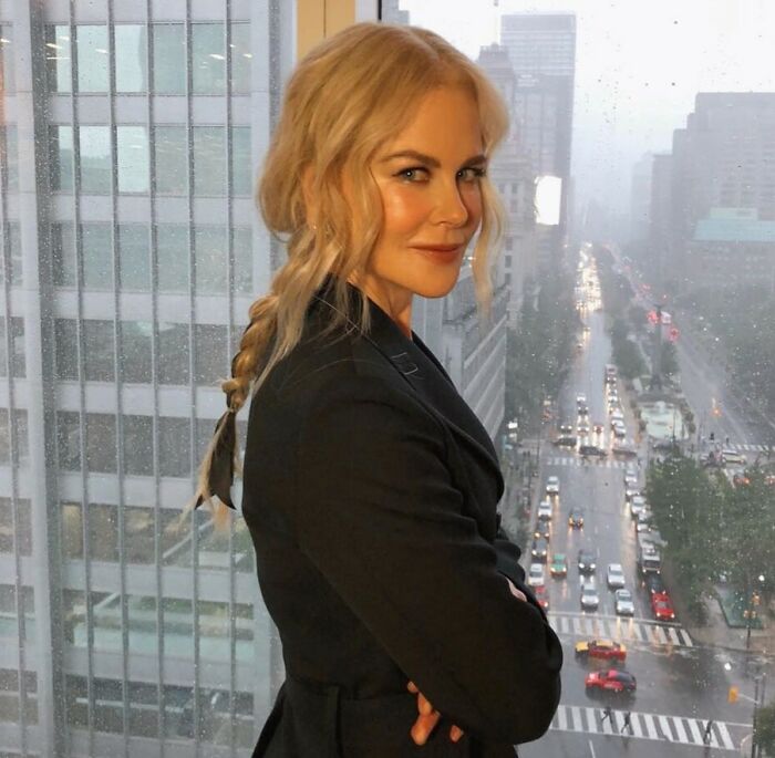 Nicole Kidman with braided hair in a black outfit, posing indoors with a cityscape and rainy street in the background.