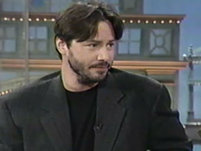 Keanu Reeves wearing a dark blazer and black shirt during a television interview in a studio setting.