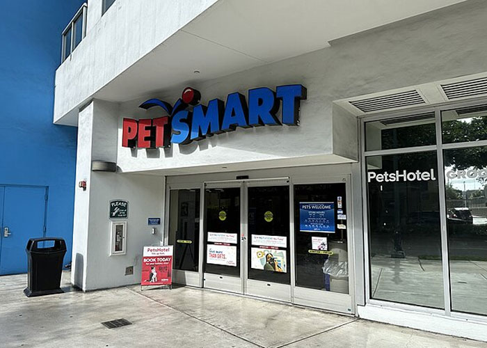 PetSmart storefront with automatic sliding doors and signs promoting PetsHotel against a sunny urban backdrop.