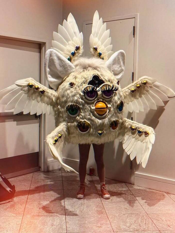 Person wearing confusing and weird costume covered in fur, multiple eyes, wings, and exaggerated facial features indoors.
