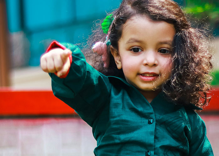 Young girl with curly hair pointing forward outdoors, capturing a moment of kids being mortified by funny antics.