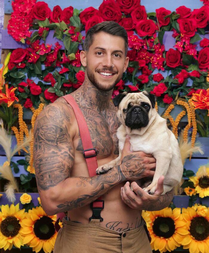 Tattooed firefighter in suspenders holding a pug, posing in front of vibrant flowers for Australian calendar photos.