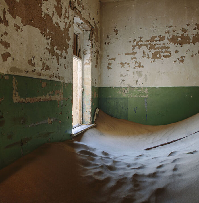 Desert sand filling an abandoned home room with peeling walls and a partially open window showing sand outside.