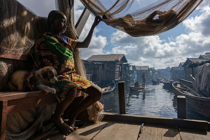 Child in colorful clothing sitting with dog inside a wooden house by water, a winning photo from the 2025 Photographer Of The Year contest