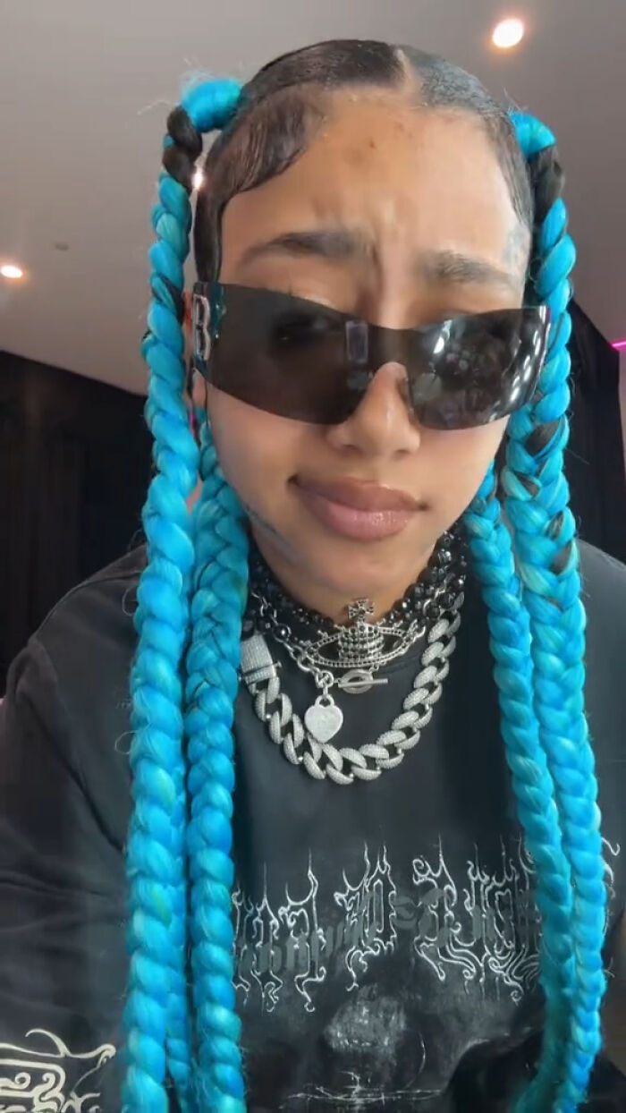 Person with blue braids wearing dark sunglasses, chains, and fake face tattoos, piercings, and grills visible on their face.