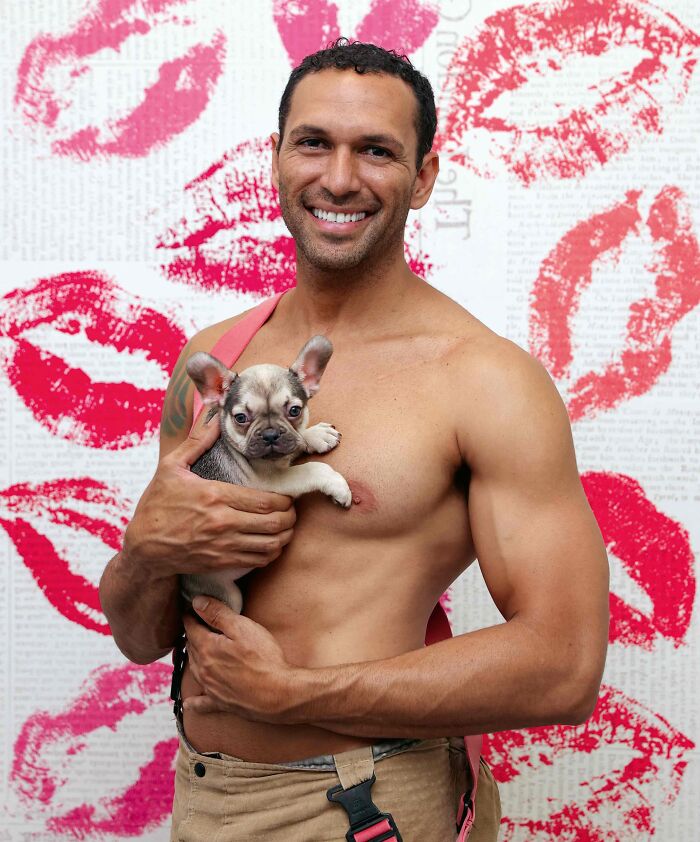 Shirtless firefighter holding an adorable dog, smiling against a backdrop with red lipstick kiss prints.