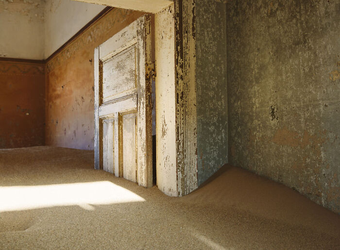 Abandoned home interior with weathered door and walls, partially buried under desert sand in a surreal sand castle scene.