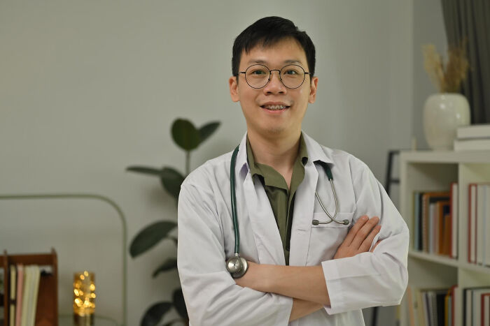 Young male doctor with glasses and stethoscope smiling confidently in a medical office, related to dementia patient care.