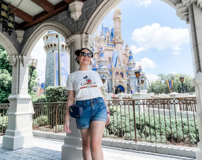 Young woman Disney superfan smiling with sunglasses near iconic castle at theme park on sunny day outdoors