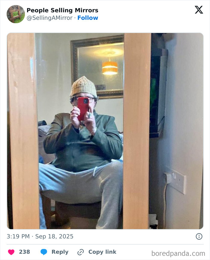 Person taking mirror selfie seated with legs wide, highlighting humorous mirror reflection in casual room setting.