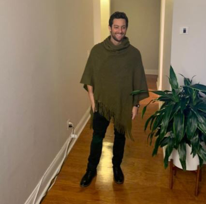 Man wearing a shawl indoors smiling while standing next to a plant in a hallway with wooden flooring.