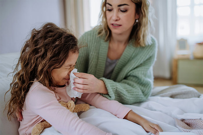 Woman caring for child, gently wiping nose with tissue, illustrating concerns about unvaccinated kid and family fallout.