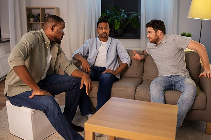 Three men having a serious conversation in a living room, illustrating wife hears husband admit with ugly woman theme.