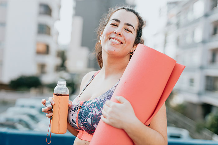 Overweight woman holding a yoga mat and water bottle at gym, reflecting on childhood trauma and overcoming challenges.