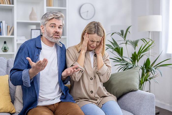 Middle-aged couple arguing on a couch with woman upset and man explaining, symbolizing golden child lost parents money crypto issue.