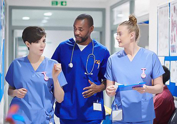 Three healthcare workers in blue scrubs discussing UK health guidance about benefits of marrying your cousin in a hospital corridor.