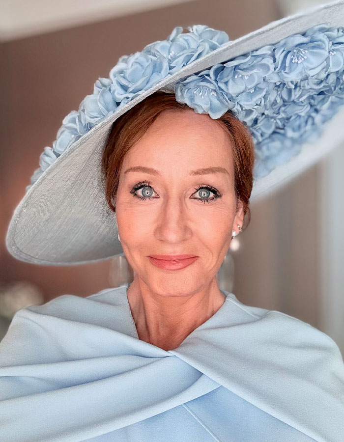 J.K. Rowling wearing a large blue floral hat and matching outfit, smiling softly in a close-up portrait.