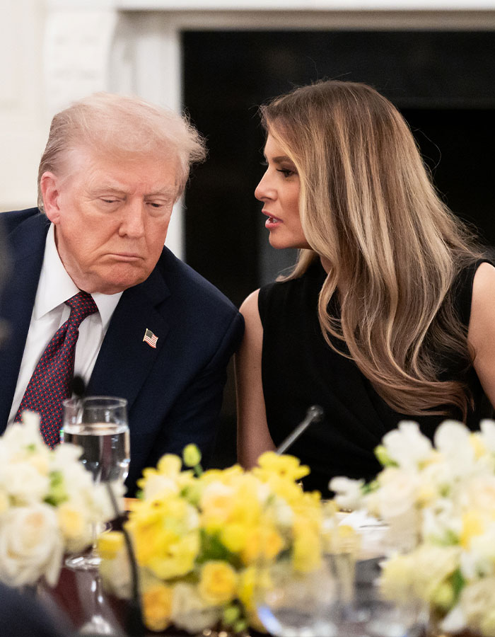 Melania Trump speaking quietly to Donald Trump during a formal event with floral table decorations nearby.
