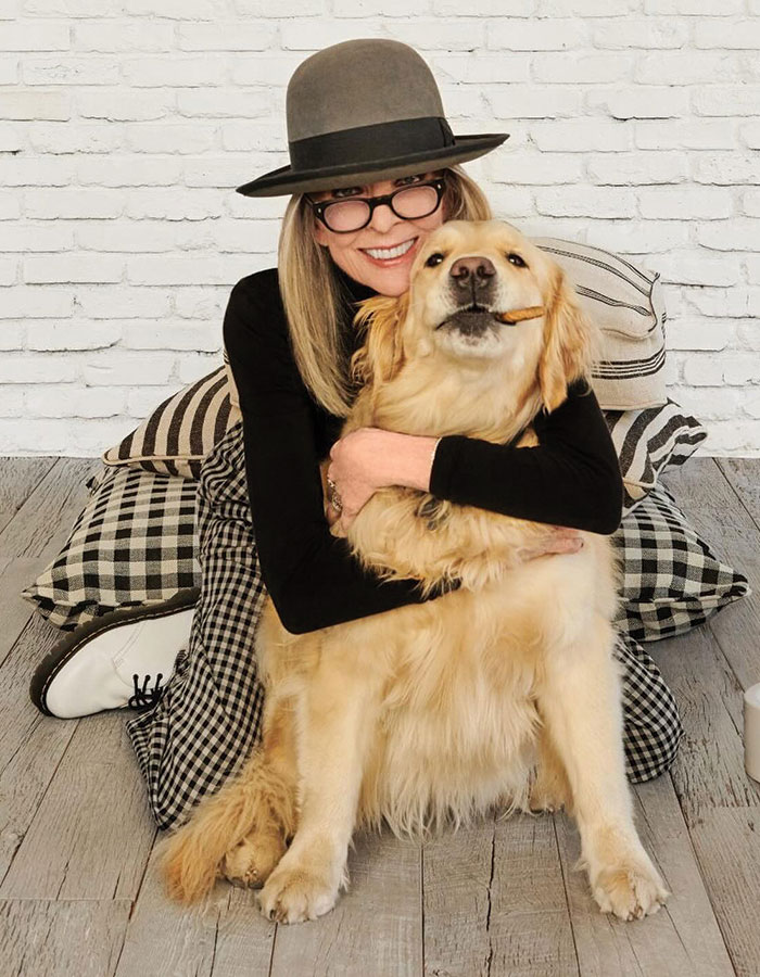 Diane Keaton sitting on floor wearing hat and glasses hugging golden retriever dog with chew stick in mouth.