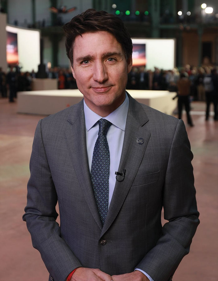 Justin Trudeau in a grey suit speaking at an indoor event with people and screens in the background.