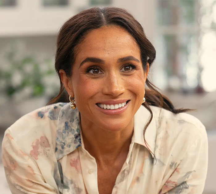 Meghan Markle smiling warmly indoors, wearing a floral blouse, related to Paris Fashion Week and Balenciaga designer.